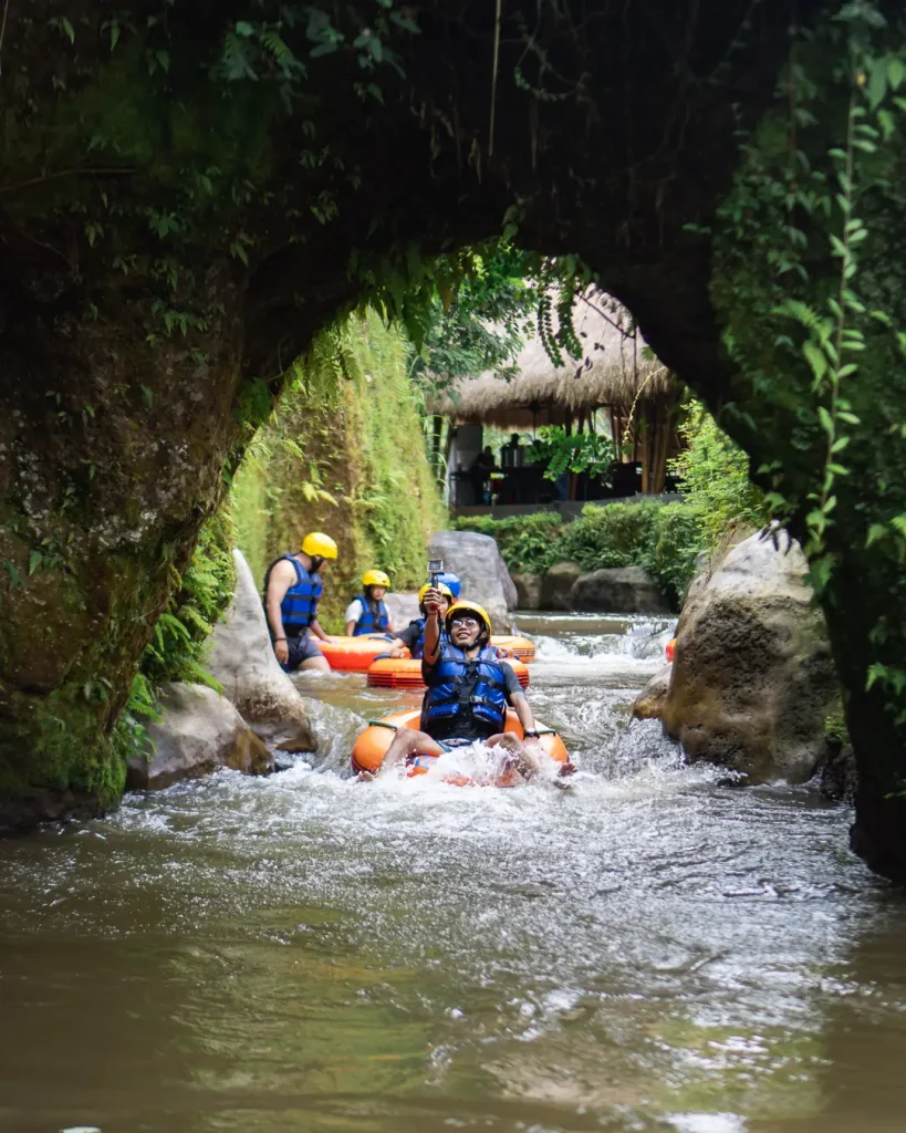 Tourists on tubes emerging from a jungle cave archway beside a thatched hut